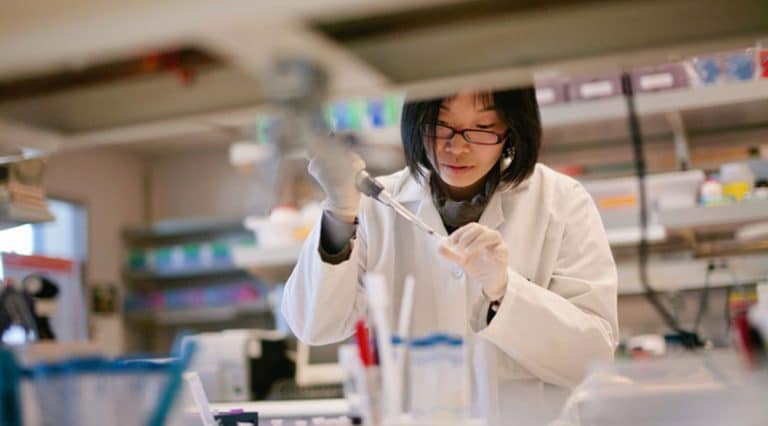 A scientist wearing a white lab coat and gloves uses a pipette in a laboratory filled with equipment and colorful supplies, conducting cancer research with adult stem cells.