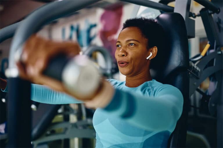 A woman wearing wireless earbuds and a blue long-sleeve top uses a chest press machine at the gym, appearing focused and determined—her drive reminiscent of the vitality often seen with younger stem cells.