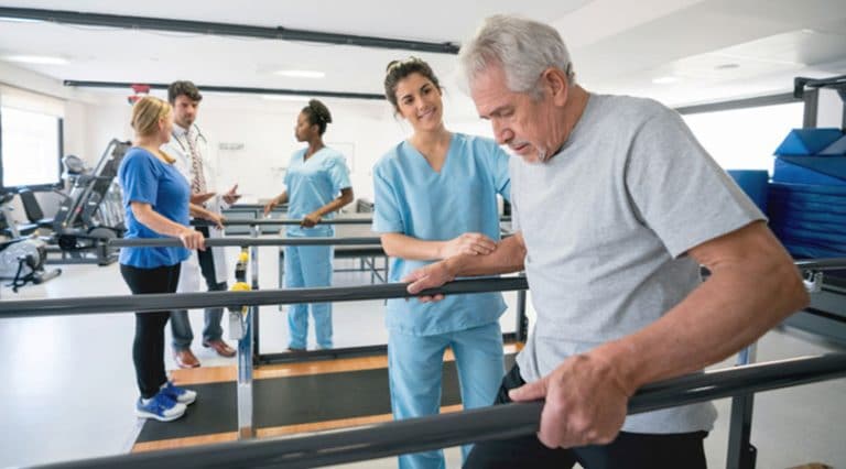 An older man uses parallel bars for support during physical therapy, assisted by a healthcare professional. In the background, other people and therapists engage in similar rehabilitation exercises in a bright gym.
