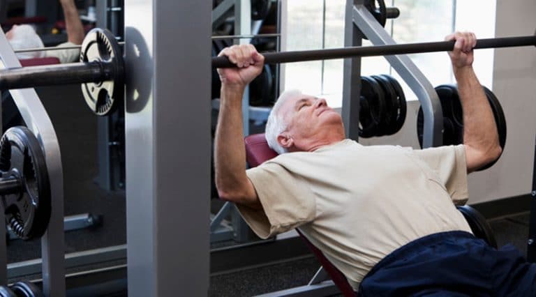An older man in a beige T-shirt is lying on a bench in a gym, lifting a barbell as part of his bench press—building strength after treatment for rotator cuff injuries. Weight plates and other gym equipment are visible in the background.