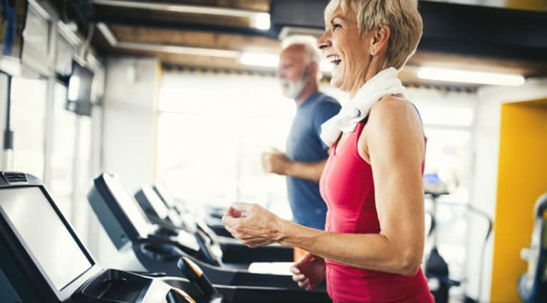 An older woman with knee osteoarthritis smiles while walking or running on a treadmill at the gym, a towel around her neck. An older man is visible in the background. Both appear to be enjoying their workout.