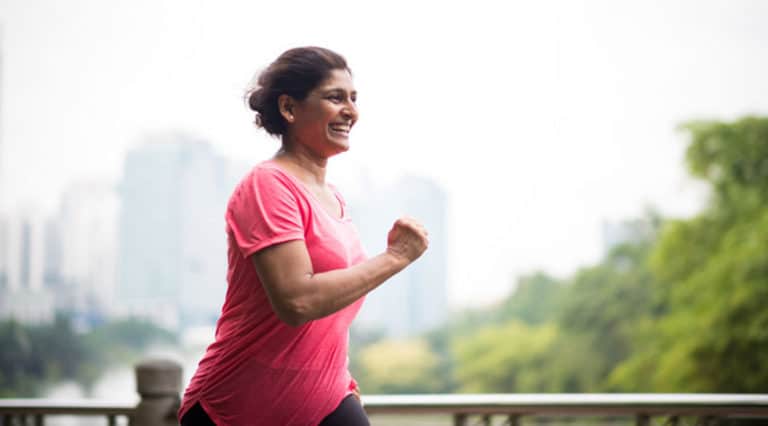 A woman in a pink shirt smiles while jogging outdoors, showing the positive impact that treated health conditions and advances in regenerative medicine can have, with blurred trees and city buildings in the background.