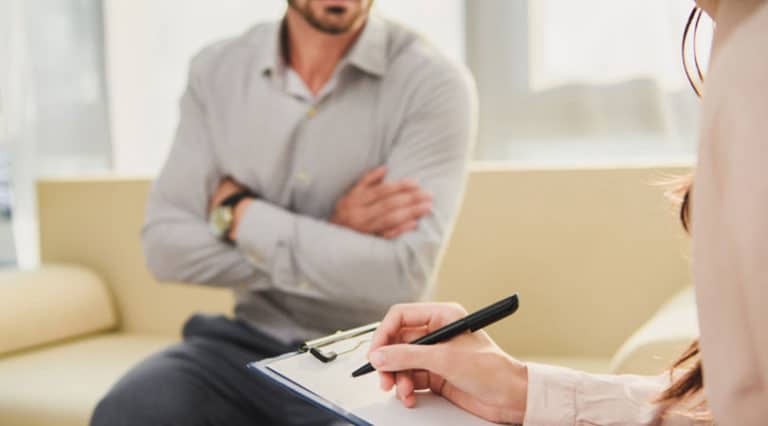 A man sits on a couch with arms crossed while another person holds a clipboard and pen, taking notes during a counseling or therapy session to discuss insurance coverage for various procedures.