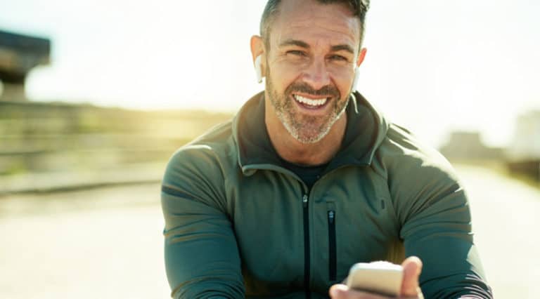 A smiling man with short hair and a beard, a highly trained medical doctor, wears earbuds and an olive-green jacket while sitting outdoors, holding a smartphone. Sunlight shines in the background.