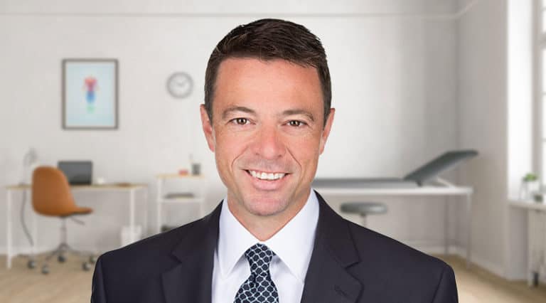 A Michigan doctor in a suit and tie smiles while standing in a bright, modern office with a desk, chair, wall art, and an examination table—specializing in advanced treatments like Cervical Intradiscal Injection.