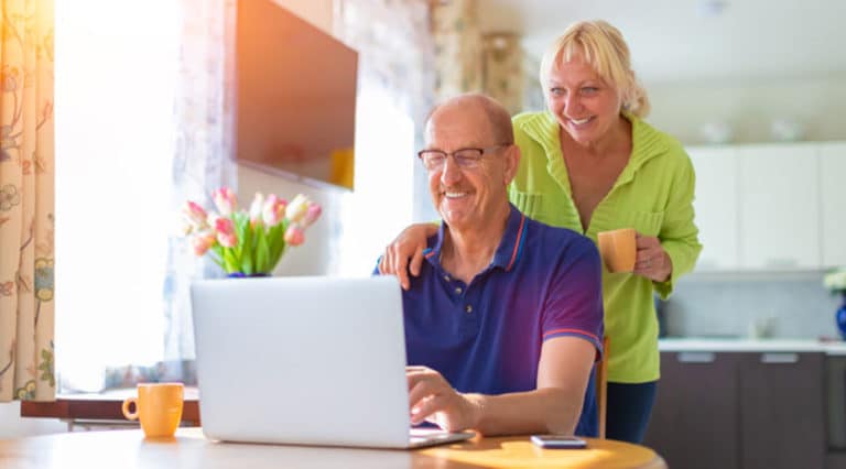 An older man sits at a table using a laptop researching PRP for hair loss while an older woman stands beside him, smiling and holding a mug. Sunlight streams through the window, and a vase of tulips is on the table.