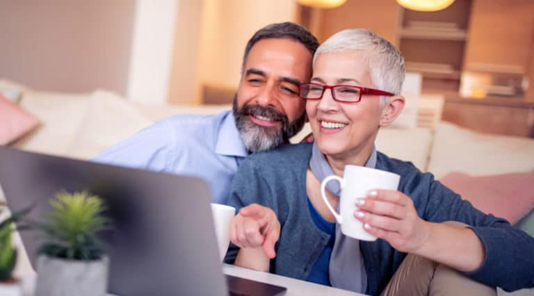 Smiling older couple sitting on a couch, looking at a laptop together. The woman holds a mug and points at the screen while the man leans in closely behind her, perhaps researching PRP injection options for sexual wellness. A small potted plant is on the table.