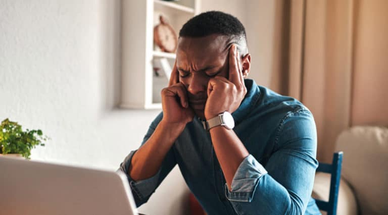 A man sitting at a desk with a laptop holds his temples and closes his eyes, appearing stressed or frustrated. He wears a denim shirt and a smartwatch, highlighting the importance of managing stress. A plant and shelves are visible in the background.