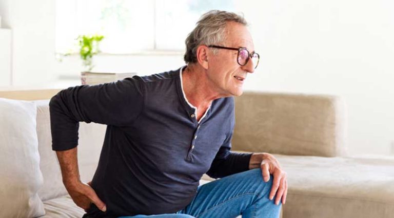 Older man wearing glasses and a dark long-sleeve shirt sits on a couch, holding his lower back with a pained expression—possibly considering back pain treatment or wondering if he’s a candidate for stem cell injections. Bright, softly lit living room in the background.