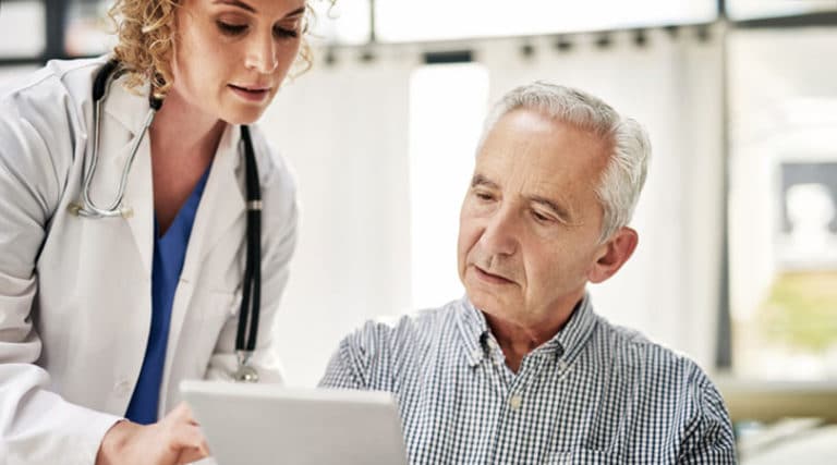 A female doctor in a white coat shows a clipboard to an older man in a checked shirt, both focused on the document during a treatment consultation, in a bright medical office setting.