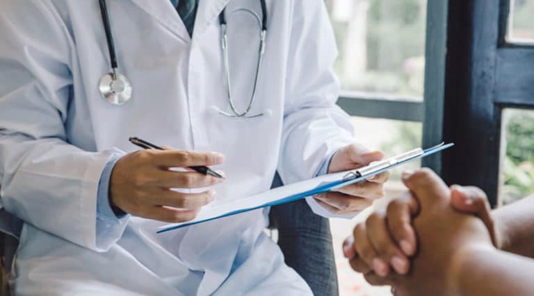A doctor in a white coat holds a clipboard and pen, discussing notes about stem cells with a patient whose hands are visible in the foreground. The scene takes place in a well-lit room with natural light from a window.
