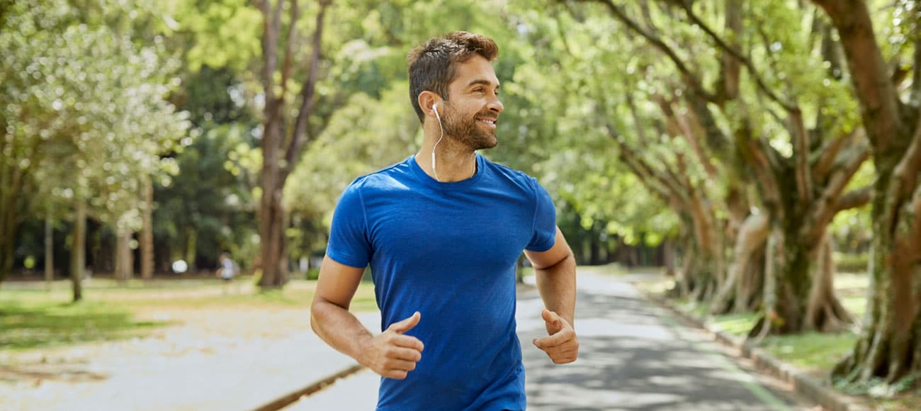 Smiling male with in-ear headphones jogging in park. Mid adult man is listening to music during summer after receiving Regenerative Medicine Near Berkley MI.