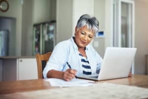 A smiling older woman with short gray hair sits at a table, using a laptop and writing on papers in a bright, modern kitchen.
