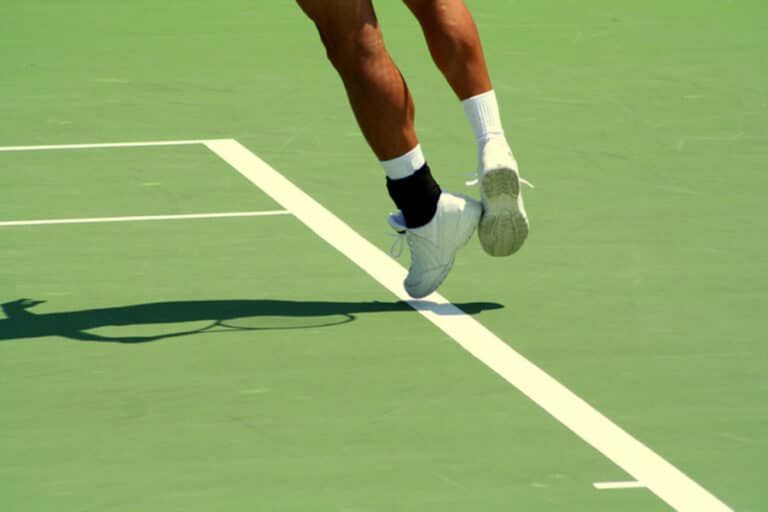 A tennis player’s lower legs and white shoes are shown off the ground as they jump on a green tennis court, casting a shadow—perhaps pushing through foot pain. White court lines are visible beneath them.
