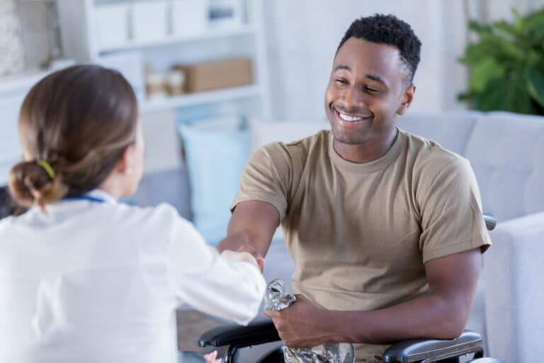 A man in a wheelchair smiles and shakes hands with a woman in a white coat, suggesting a friendly conversation about knee treatments or support session in a bright, comfortable room.