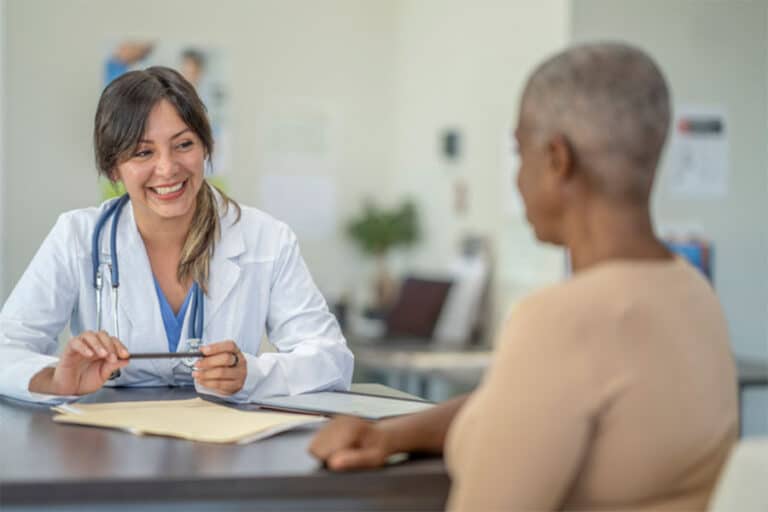 A smiling female doctor in a white coat sits at a desk, discussing new treatment options with a patient. The doctor holds a pen and folder, while the patient listens attentively in the bright medical office.
