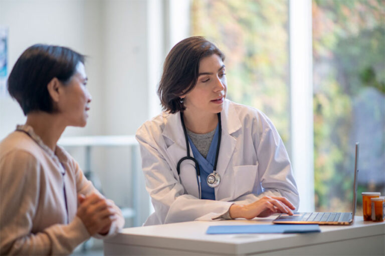A doctor in a white coat, with a stethoscope, sits at a desk and explains ways to delay diabetic neuropathy on a laptop to a patient in a light sweater. They appear to be having a serious conversation in a bright office.