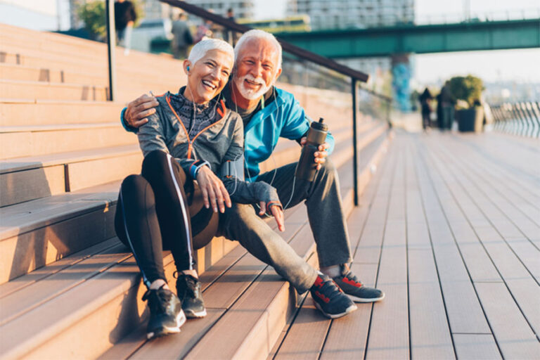 An older couple in athletic wear sits on outdoor steps, smiling and embracing after exercise. The man holds a water bottle, celebrating recovery from an Achilles tendon injury through stem cell therapy under the warm, cheerful sun.