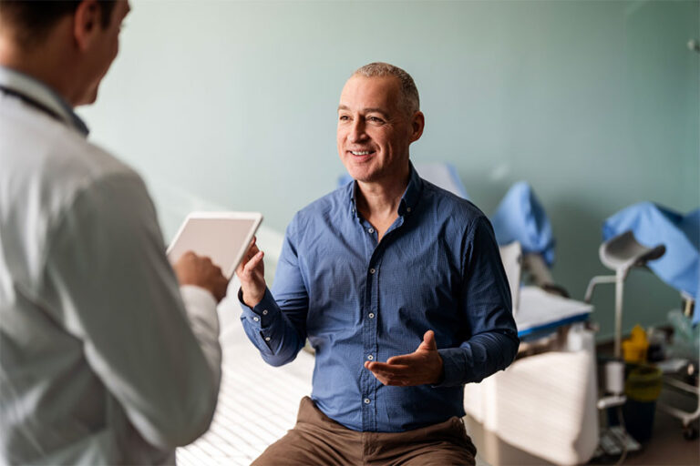 A middle-aged man sits on an exam table, smiling and talking with a healthcare professional about peripheral neuropathy treatment options as the provider reviews information on a tablet. Medical equipment is visible in the background.