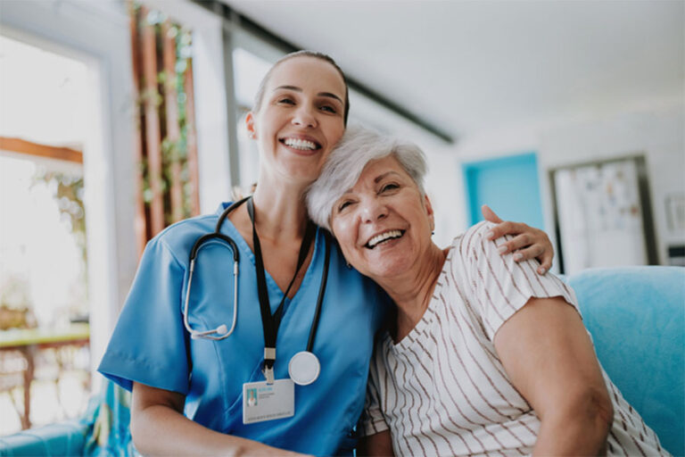 A smiling nurse in blue scrubs with a stethoscope embraces an older woman with gray hair, who is also smiling. They are sitting together in a bright, cheerful room at the Michigan Center for Regenerative Medicine.