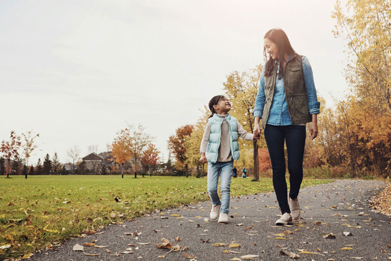 A woman and a young girl walk hand in hand on a paved path in a park with autumn trees and fallen leaves, smiling at each other under a bright sky—a serene moment reflecting the vitality of advanced pelvic floor health.