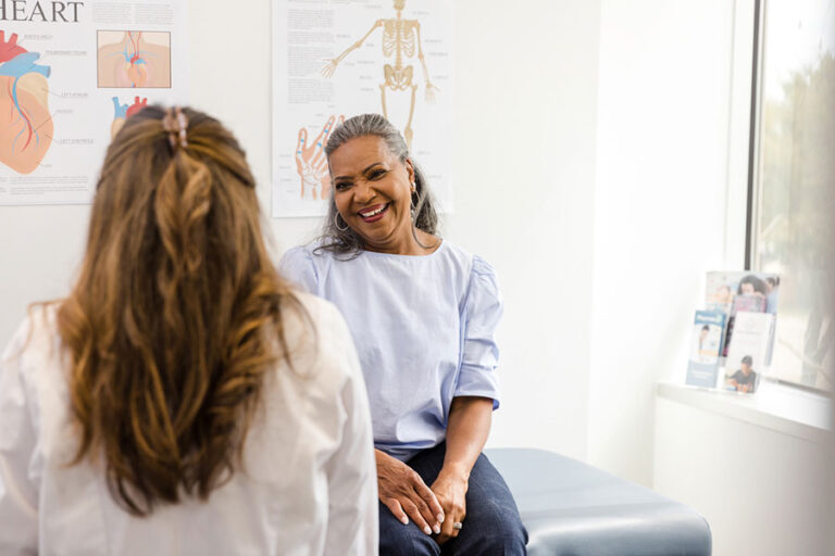 A smiling woman sits on an exam table in a doctor's office, discussing pelvic floor exercises with a healthcare professional. Medical posters line the wall, and sunlight streams through a nearby window.