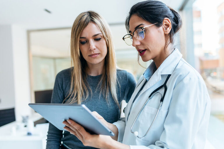 A doctor in a white coat with a stethoscope shows a digital tablet to a woman in a gray sweater, discussing O-Shot benefits in a bright, modern office.