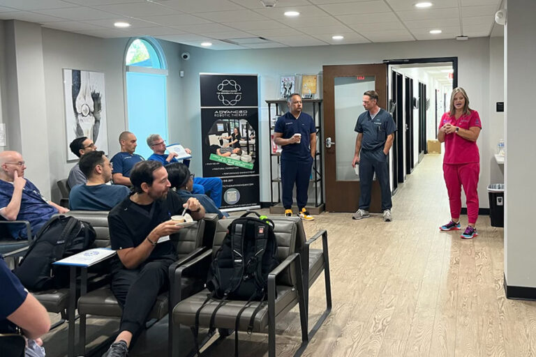 A group of people sit in chairs, facing three standing speakers in a bright, modern room with wood floors and medical posters. At this Midwest Training Hub, one speaker in pink scrubs discusses Advanced Orthobiologic Techniques with the audience.
