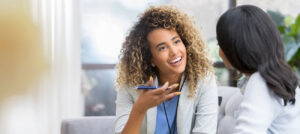 Two women are sitting and talking indoors in Rochester MI. One woman with curly hair smiles while holding a smartphone, discussing skincare tips and PRP treatment with the other woman, who has dark straight hair. Natural light streams through the windows behind them.