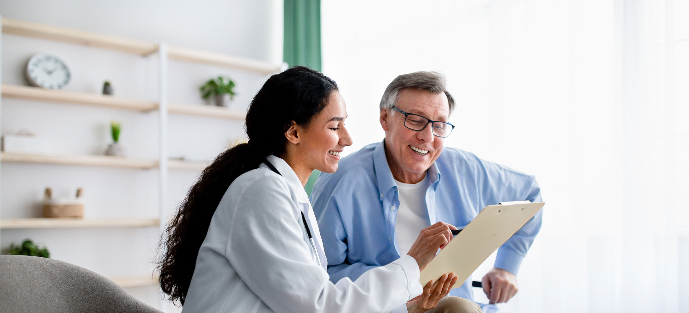 A female doctor in a white coat smiles while holding a clipboard and discussing EMG testing with an older man wearing glasses and a blue shirt in a bright, modern Dearborn MI office.