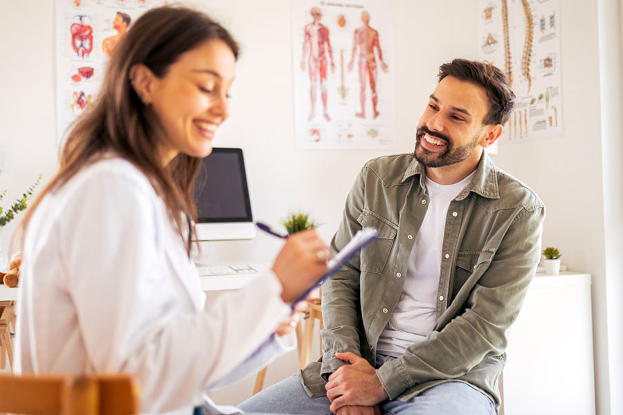 A smiling man sits and talks with a female doctor taking notes on a clipboard in a bright medical office with anatomical charts on the walls.