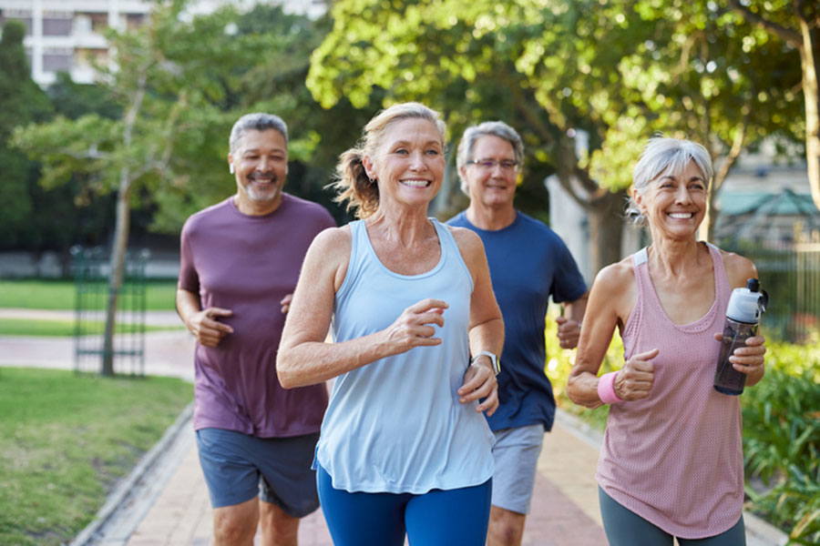 Four smiling older adults jog together on a park path surrounded by trees. Two women are in front, one holding a water bottle, with two men running behind them, all wearing athletic clothing on a sunny day.