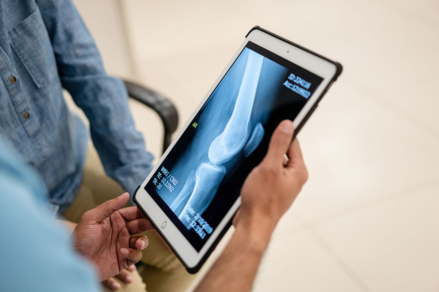 A person holds a tablet displaying an X-ray image of a human elbow, while another individual sits nearby, suggesting a medical consultation or review of the X-ray results.