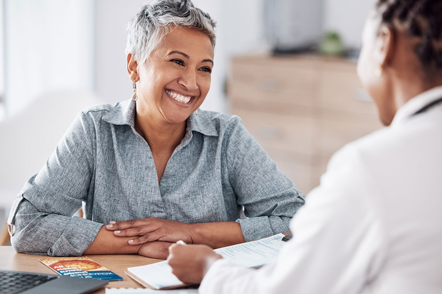 A smiling woman with short gray hair sits at a desk, talking to a healthcare professional holding a clipboard. Pamphlets and documents are on the desk in a bright, modern office setting.