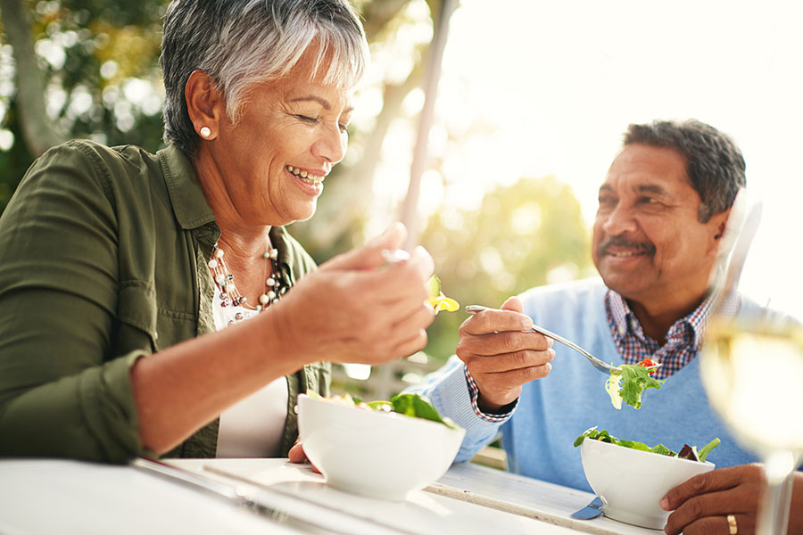 An older couple sits outdoors at a table, smiling and enjoying fresh salads together on a sunny day. The woman is holding a fork with greens; the man looks at her, also holding a forkful of salad.