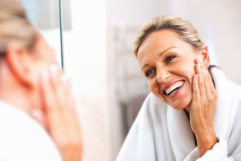 A woman in a white bathrobe stands in front of a mirror, smiling and gently touching her cheek with one hand, appearing happy and relaxed in a bright bathroom after using transforming skincare.
