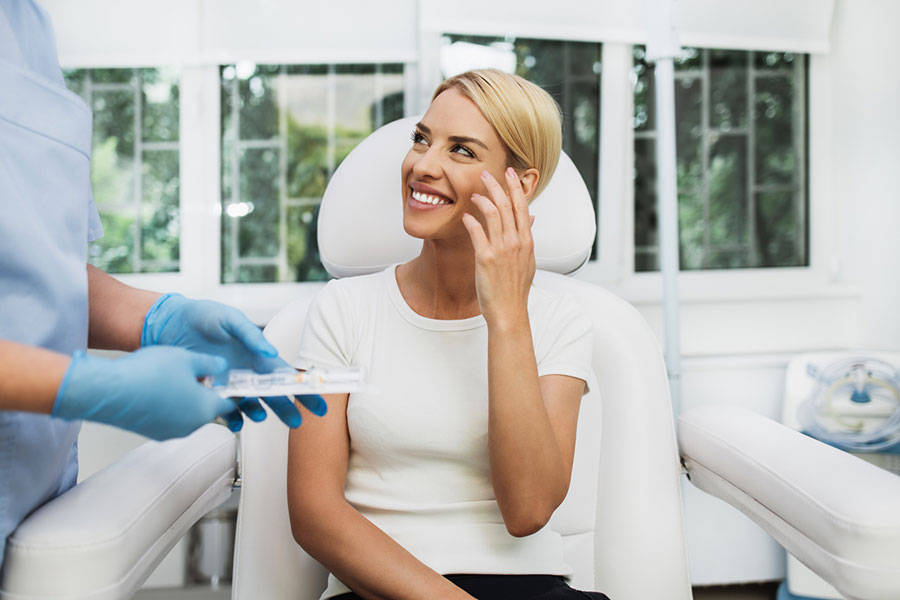 A woman sits in a medical chair, smiling and touching her face, while a healthcare professional wearing blue gloves stands nearby holding a tray—highlighting the promise of skincare and visible skin transformation in a bright, modern clinic.