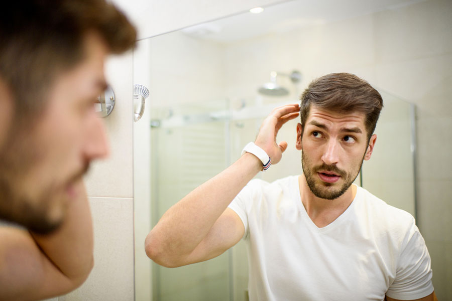 A man with short brown hair and a beard looks at himself in a bathroom mirror, touching his hair with one hand. He appears to be inspecting for thinning or hair loss while wearing a white t-shirt and a white wristwatch.