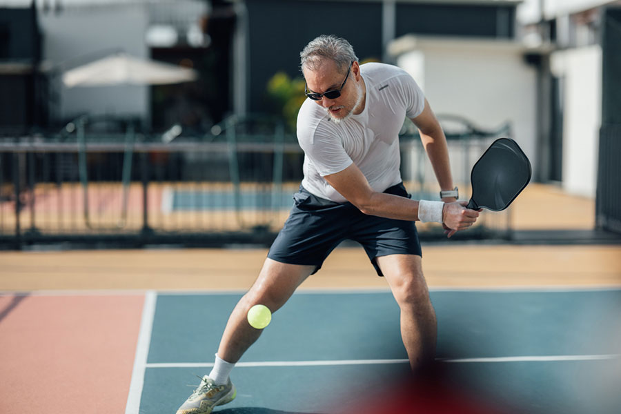 A man wearing sunglasses and athletic clothes, possibly recovering from plastic surgery, plays pickleball on an outdoor court, preparing to hit a yellow ball with his paddle.