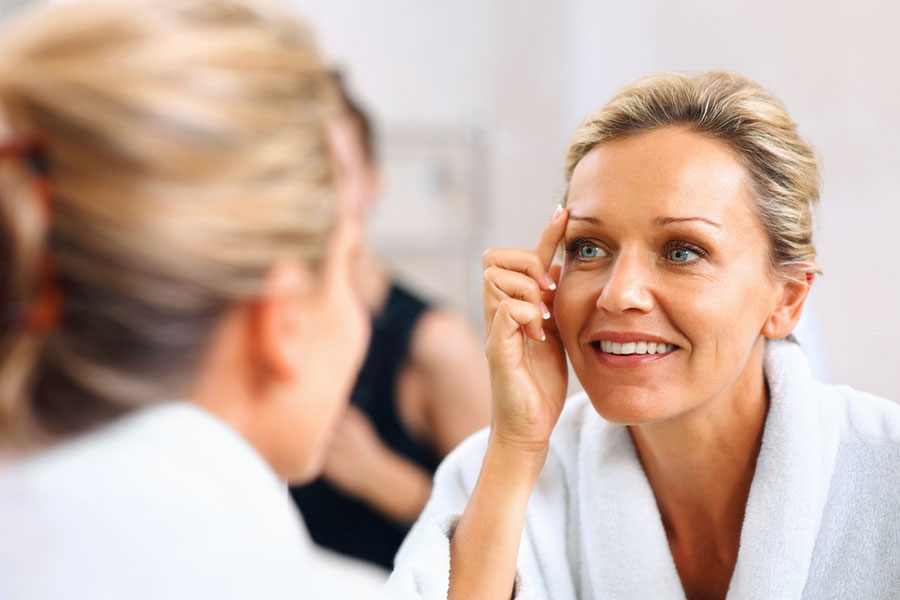 A woman in a white bathrobe looks at herself in the mirror, smiling and touching the skin near her eye with her finger—a smart alternative to plastic surgery. The background is softly blurred.