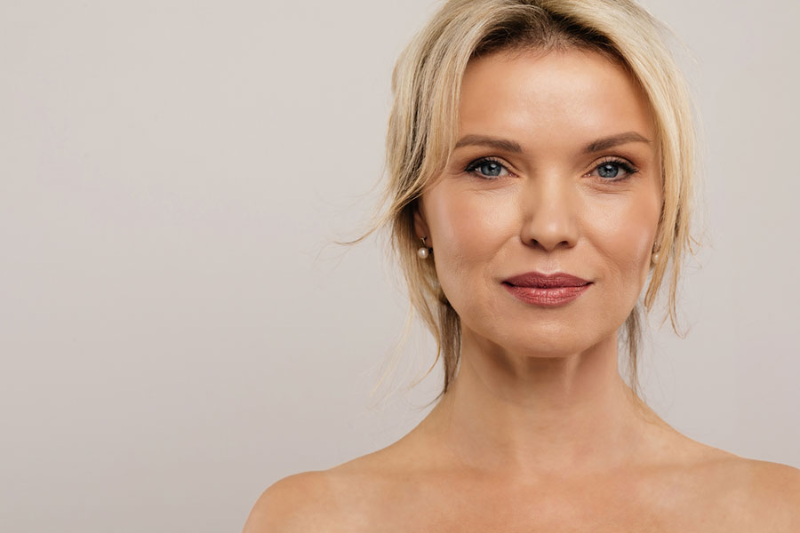Portrait of a woman with blonde hair, light eyes, and natural makeup, looking confidently at the camera against a plain, light background. She has a calm expression and bare shoulders, exemplifying the smart alternative to traditional plastic surgery.