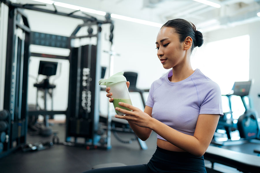 A woman in a light purple workout top sits in a gym, holding a green smoothie in a shaker bottle—an essential part of her sports nutrition routine—while exercise equipment fills the background, highlighting her dedication to athletic performance.