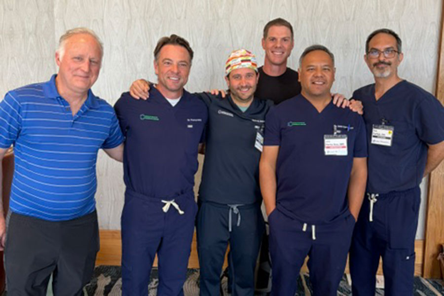 Six men, five in medical scrubs and one in a blue polo, stand together indoors smiling at the camera. The group appears friendly and relaxed, suggesting a team or colleagues gathering—perhaps attending a regenerative medicine conference in Michigan.