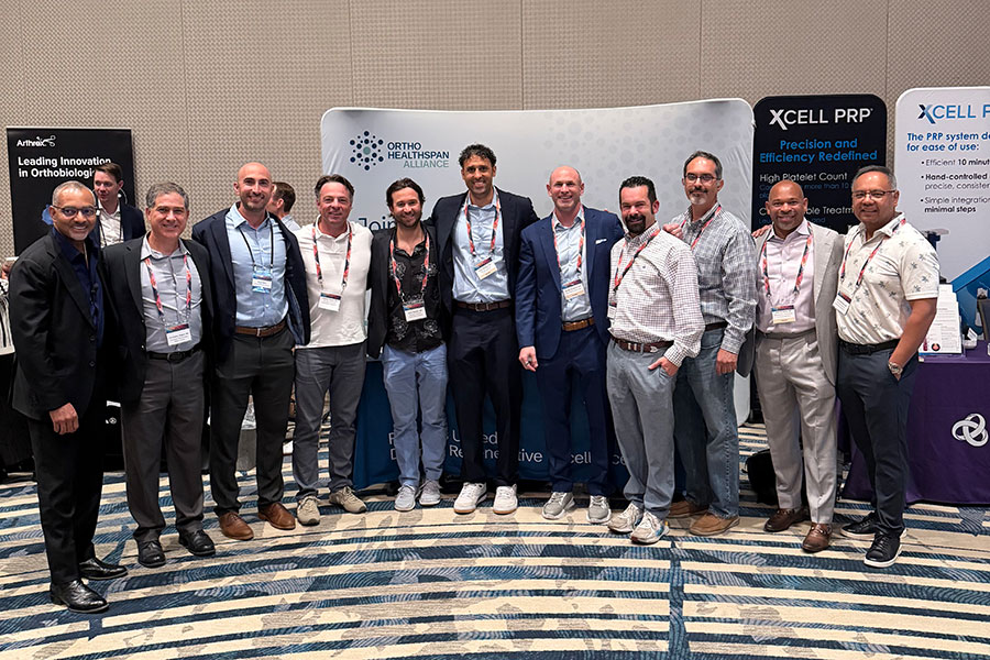 Eleven men stand in a row, smiling, in front of exhibitor banners at a conference in Michigan. They wear business or business-casual attire and conference badges. The background shows a gray wall and displays about medical technology and regenerative medicine.
