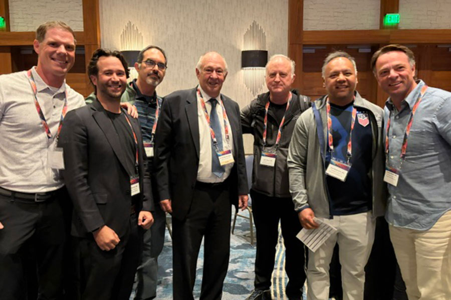 Seven men wearing conference badges stand together, smiling for a group photo at the Top Regenerative Medicine Conference in a conference room with wood paneling, carpeted floor, and decorative wall designs visible in the background.