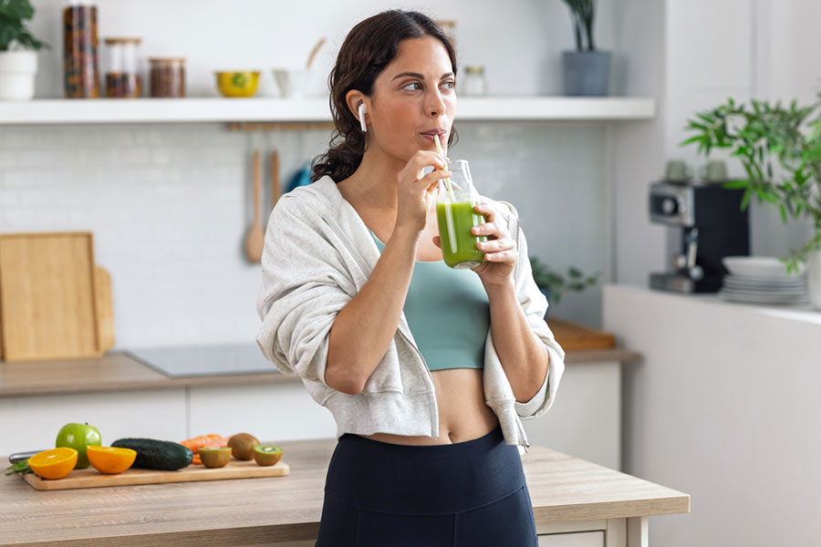 Woman in workout clothes drinks a green smoothie from a jar with a straw in a kitchen, finding balance in her morning routine. Fruits and vegetables are on the counter, supporting her efforts to reduce inflammation. She wears wireless earbuds and looks to the side, smiling.