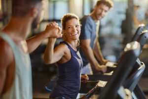 A woman smiles and fist-bumps a man while using a treadmill at the gym, celebrating their dedication to heal the body through movement. Another man, also on a treadmill, looks on and smiles in the background.