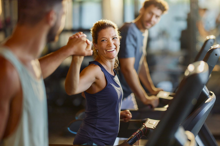 A woman smiles and fist-bumps a man while using a treadmill at the gym, celebrating their dedication to heal the body through movement. Another man, also on a treadmill, looks on and smiles in the background.