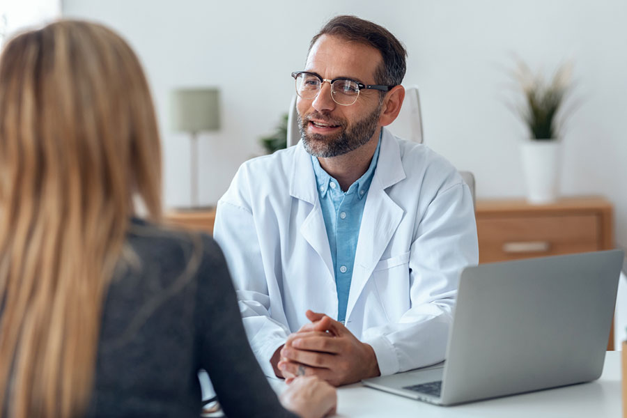 A male doctor wearing glasses and a white coat sits at a desk with a laptop, talking to a female patient. The setting appears to be a medical office with plants and a lamp in the background, creating an atmosphere that emphasizes natural healing and comfort.