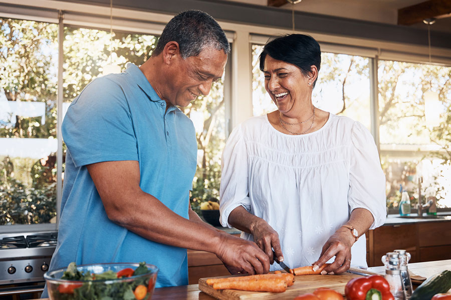 A smiling couple stands in a bright kitchen, chopping vegetables together on a wooden cutting board. Fresh produce, including tomatoes and greens known for their natural healing properties, are on the counter. Sunlight streams in through large windows behind them.